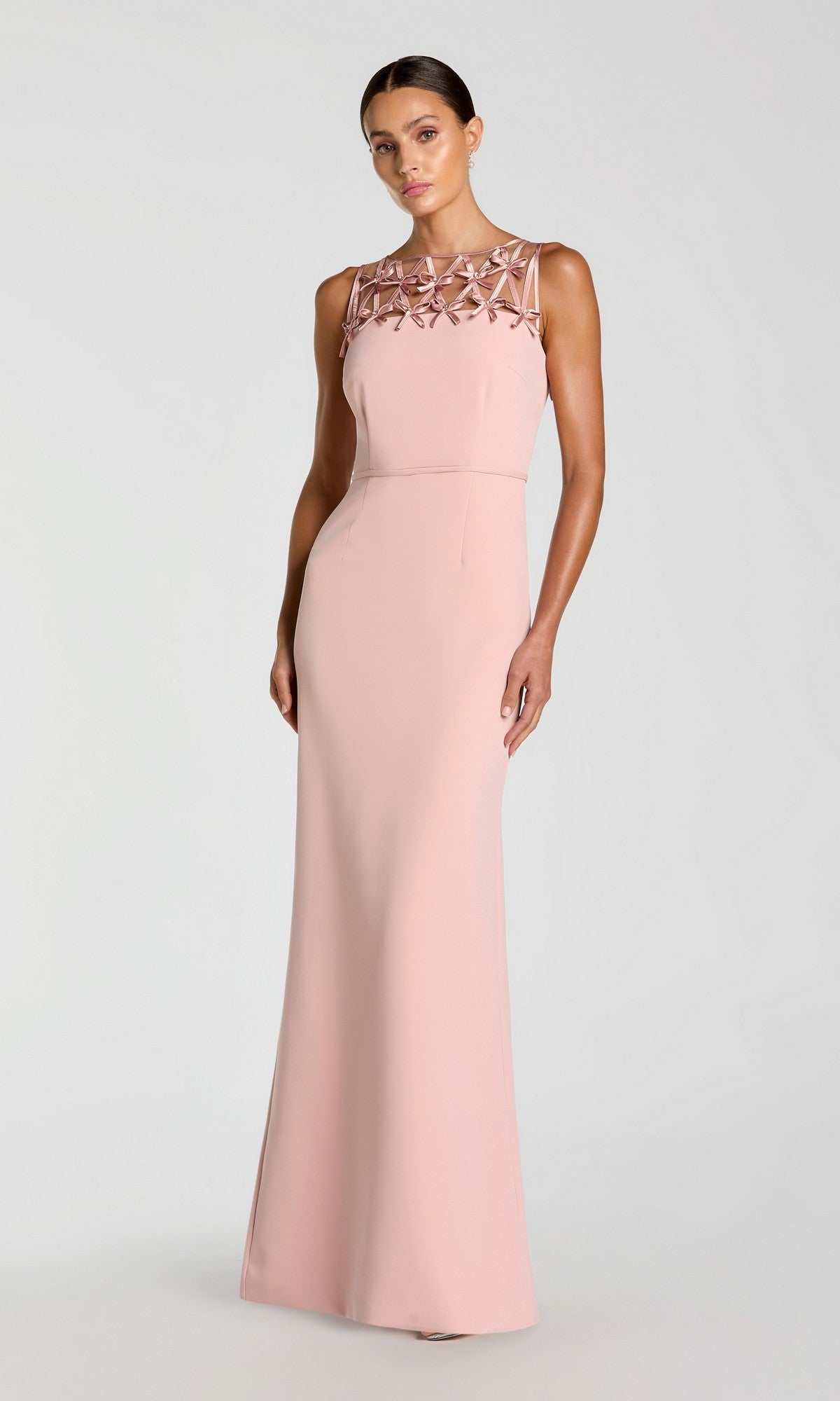 A woman wearing a sleeveless, floor-length black Mac Duggal 2295 formal dress with a bow-neck detail, standing against a plain light background.